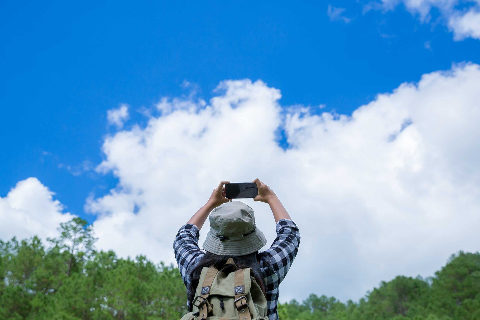 Hiker from behind photographing a vibrant blue sky with fluffy white clouds.