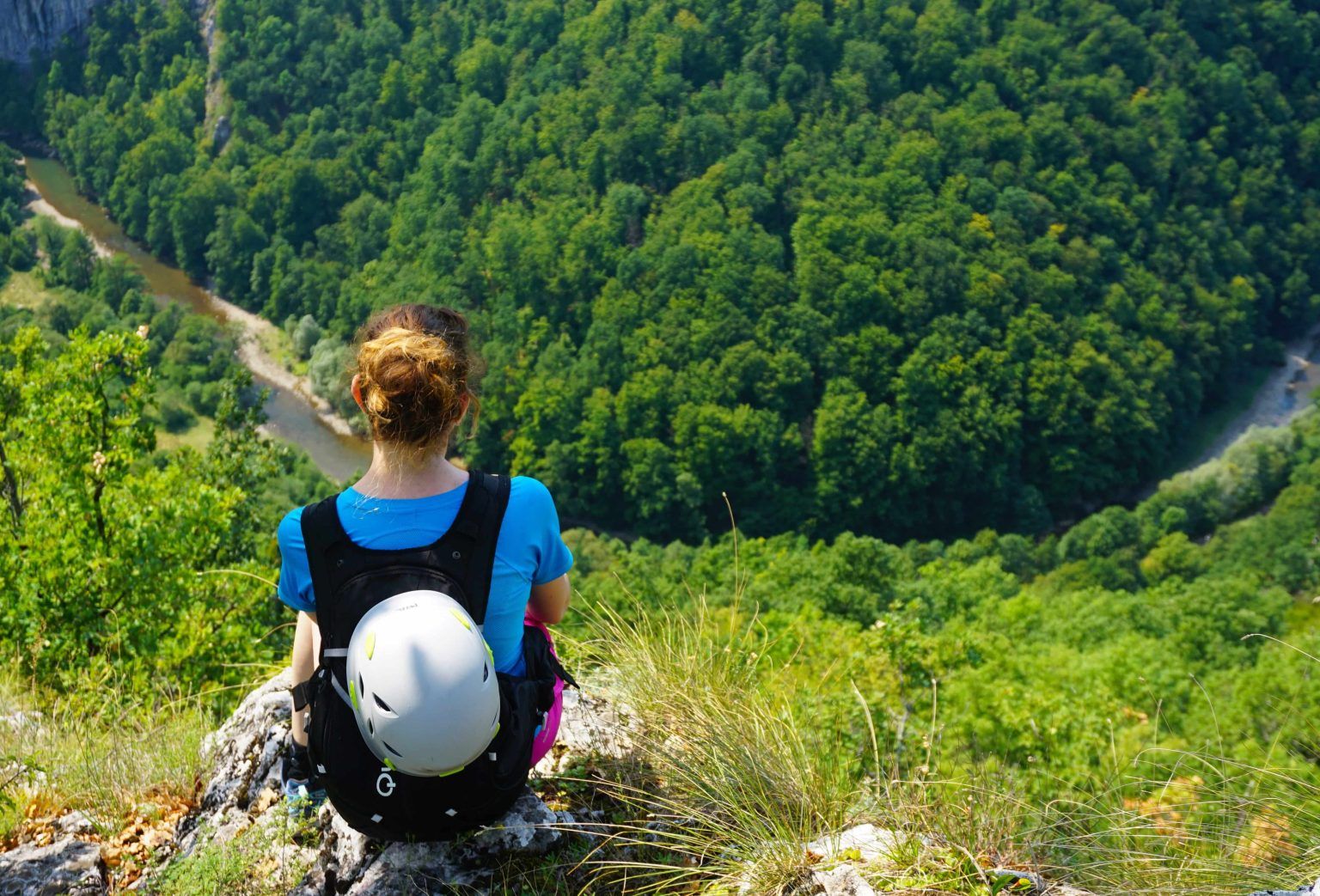 Person sitting on a cliff overlooking a vast green forest and winding river.
