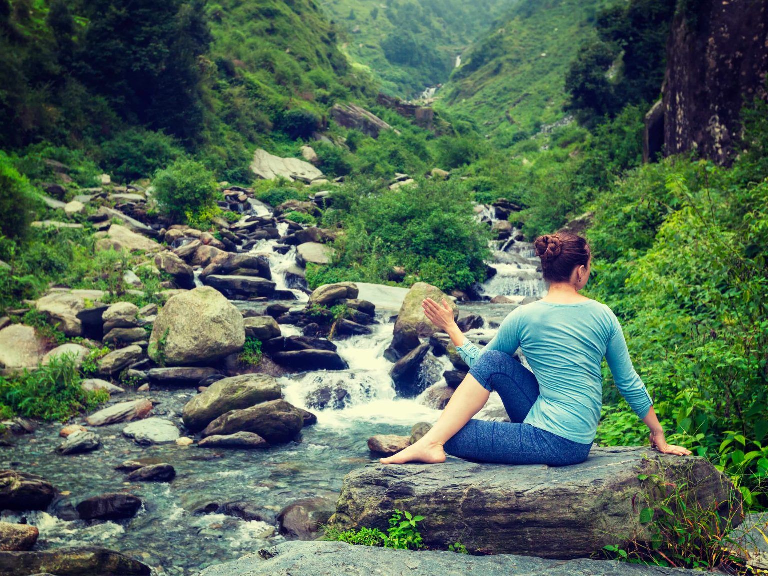 Person doing yoga on a rock by a clear river in a lush green mountain valley.