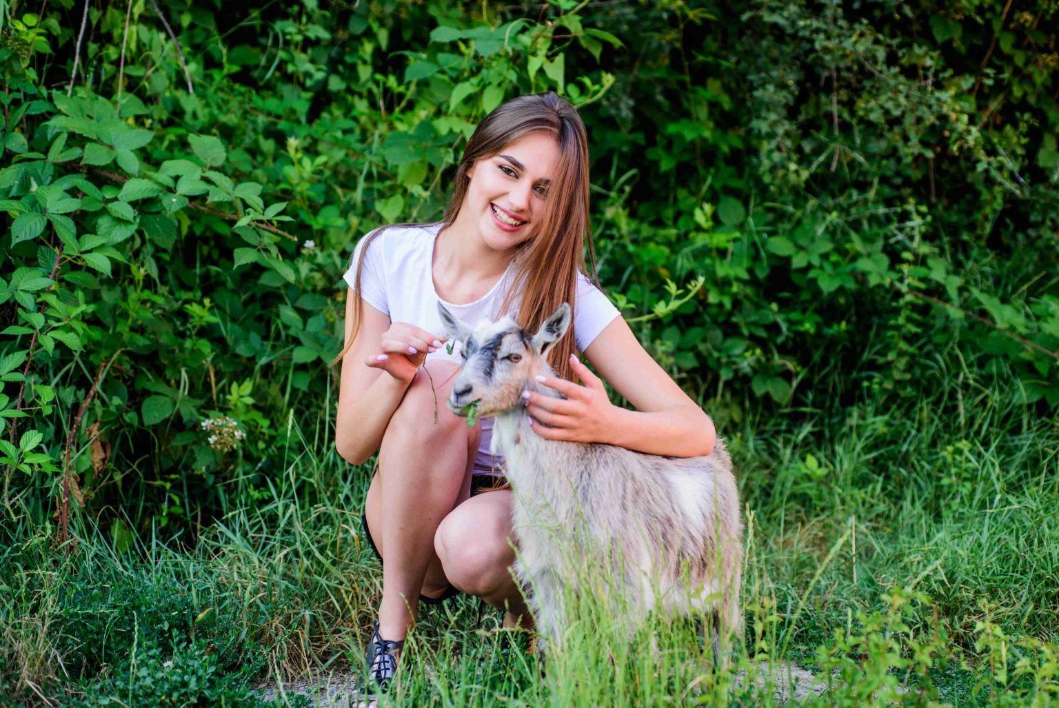 Young woman smiling and petting a small goat in a green, natural setting.