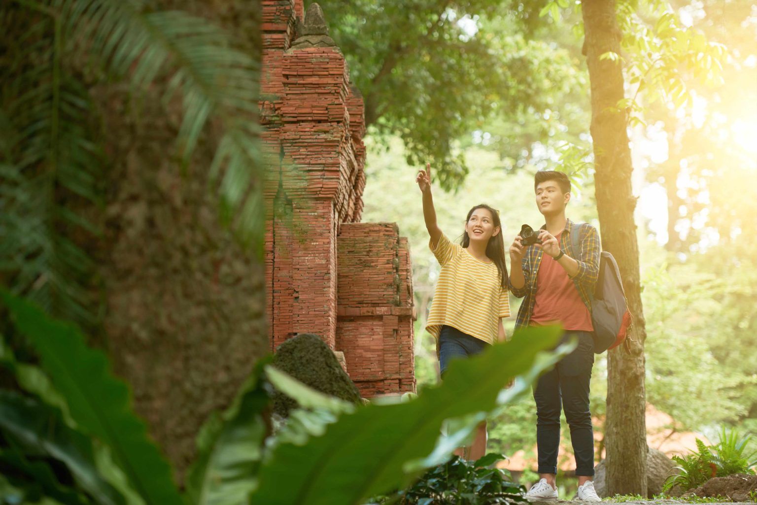 Two young tourists explore ancient brick ruins with lush greenery and bright sunlight.